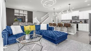 Un salon moderne avec un canapé d’angle bleu, une table basse en verre et un coin cuisine doté de meubles blancs et d’appareils électroménagers en acier inoxydable.