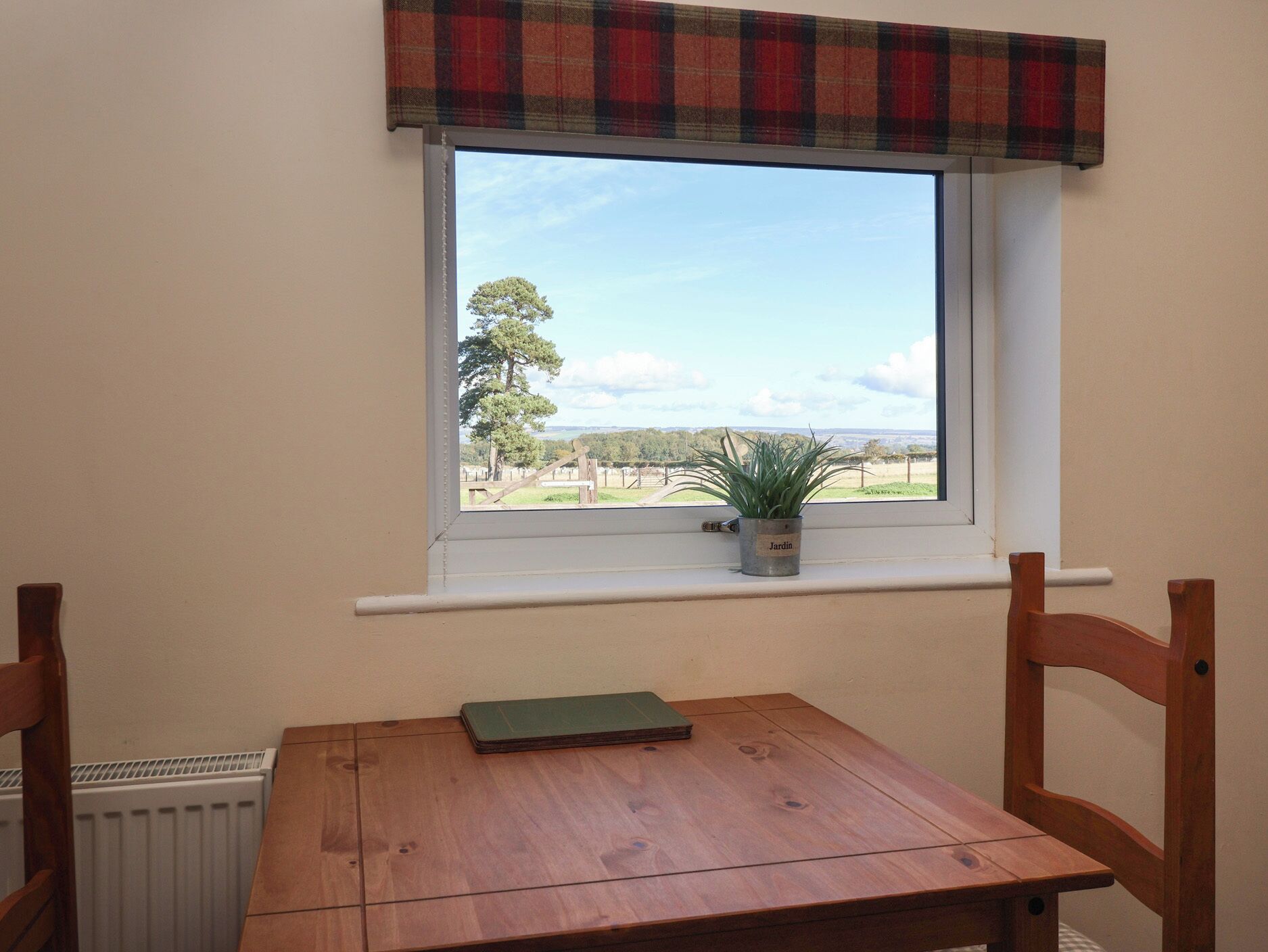 Una habitación con ventana que da a un paisaje rural, una pequeña mesa con un libro y una planta en maceta en el alféizar.