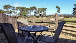 Une table ronde en métal, entourée de quatre chaises, posée sur un sol de gravier, et offrant une vue sur une zone délimitée par une clôture, avec des arbres et un paysage lointain.
