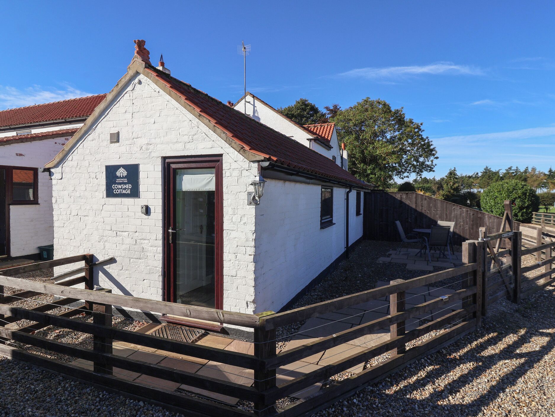 A white cottage with a red-tiled roof, a wooden fence, and a sign indicating it's part of Hartwood Holiday Park.