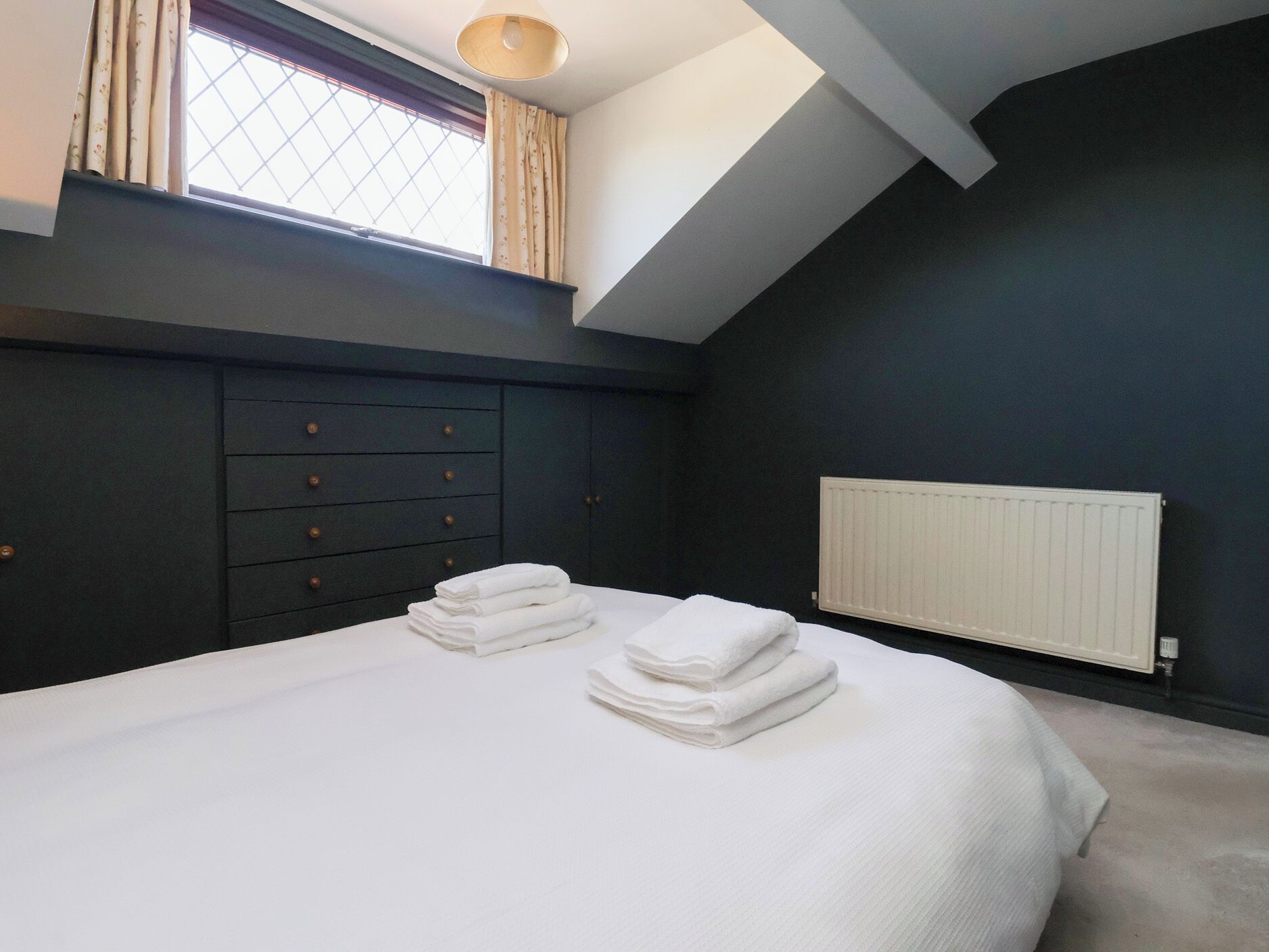 A bedroom with a large bed, a dark wall, a window with curtains, a radiator, and a ceiling light fixture.