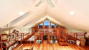 Une pièce spacieuse située sous les combles, avec un plancher en bois, un ventilateur de plafond et une vue sur l’extérieur grâce à une grande fenêtre.