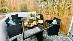 Un'area patio con tavolo in vetro, sedie in vimini e una selezione di prodotti per la colazione, tra cui pane, frutta e succo di frutta.