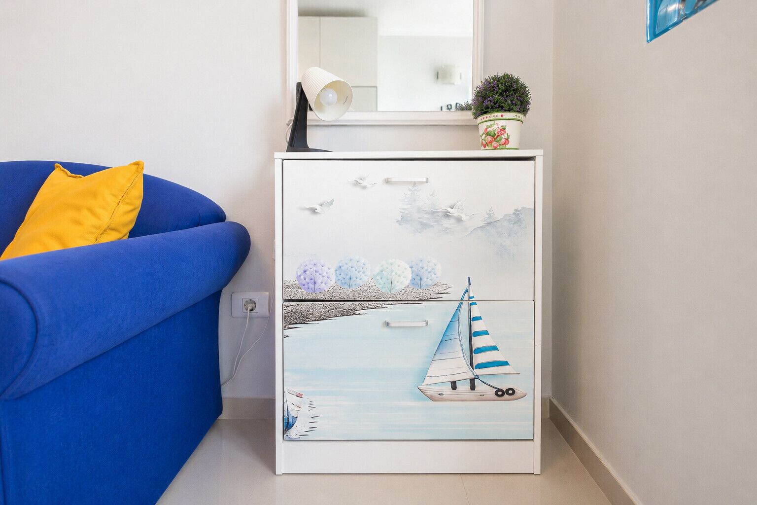 A room with a blue sofa, a white cabinet with a painting of a sailboat, and a small potted plant on top.