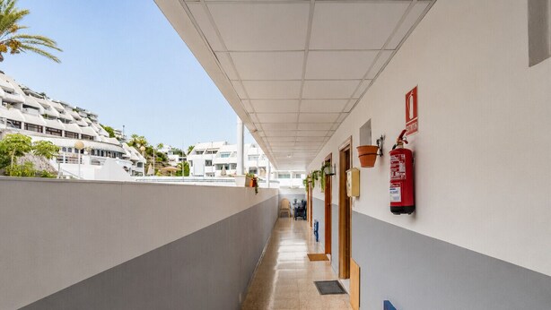 A corridor with a fire extinguisher mounted on the wall, a blue shutter, and a view of buildings in the background. - Sun Port Ocean Views - Renovated & Pool 1 (Puerto Rico)