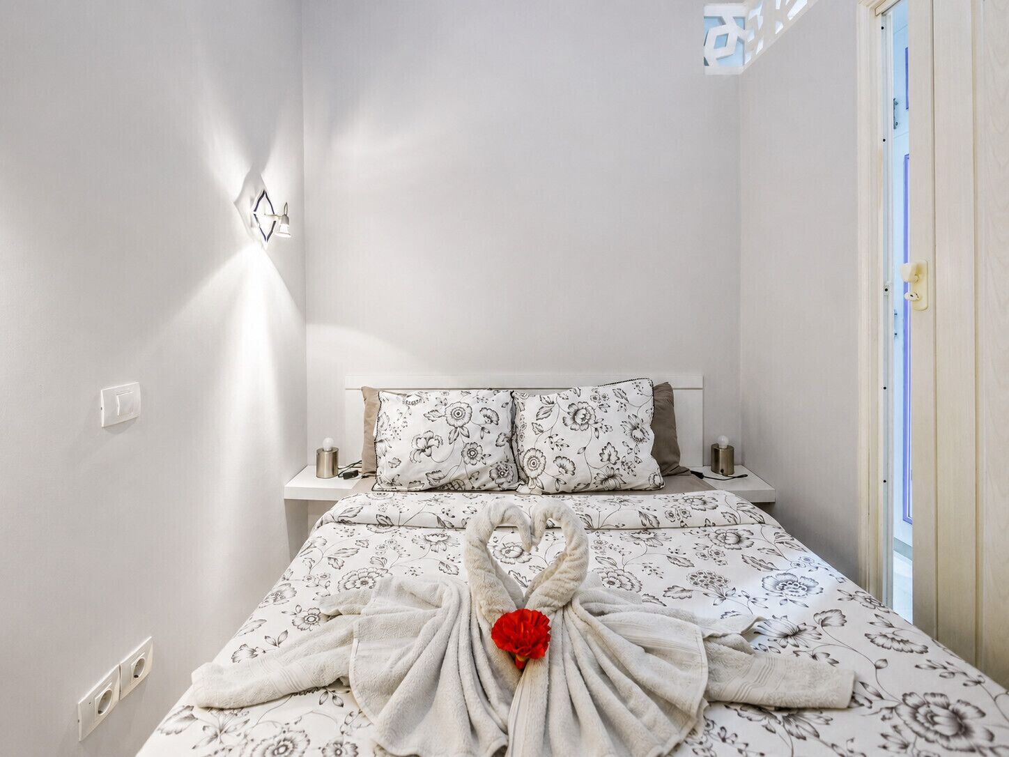 A hotel room with a bed, a nightstand, a wall-mounted light fixture, and a door.