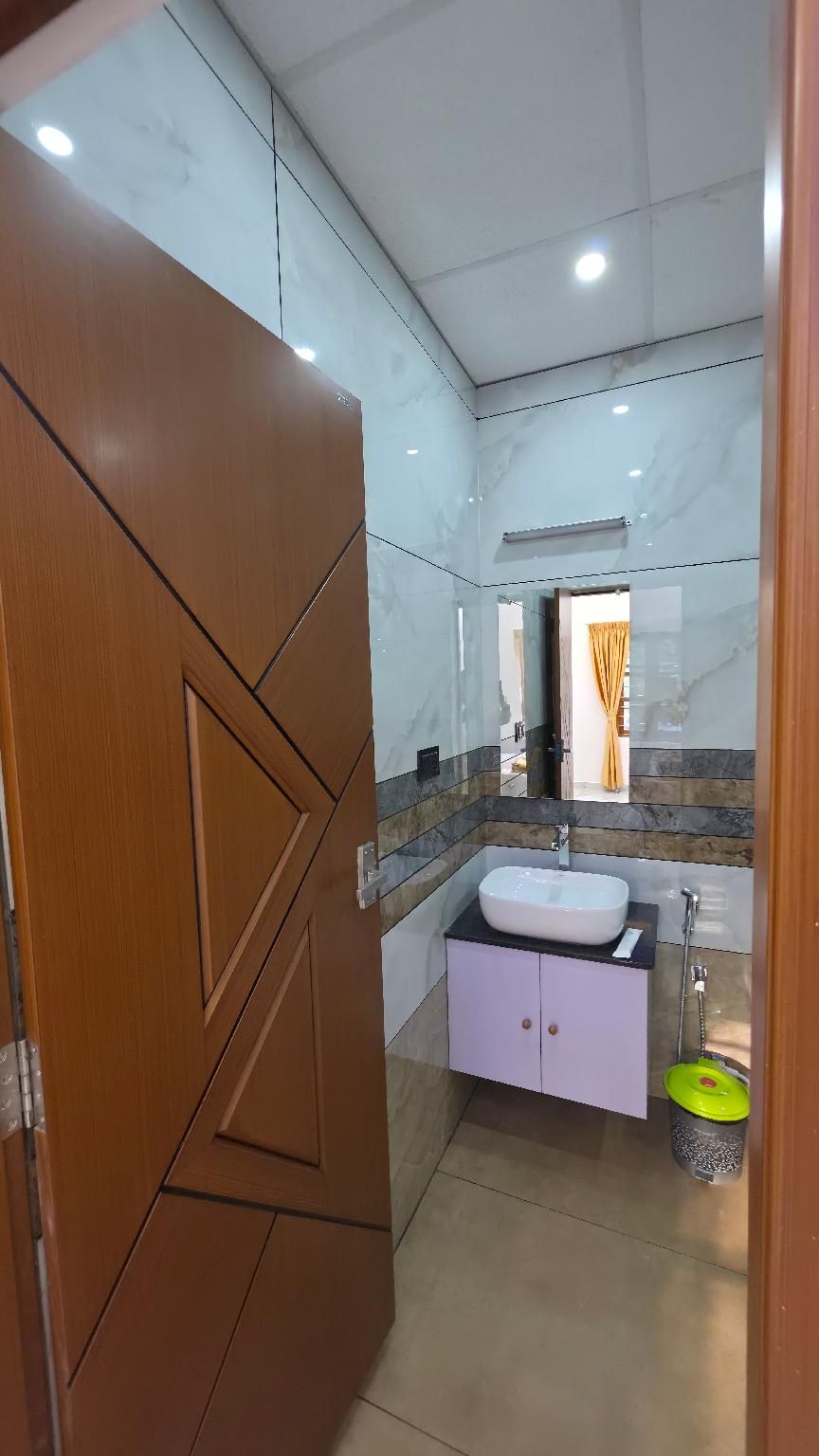 Un baño con piso de mármol, puerta de madera, lavamanos blanco y un cubo amarillo.