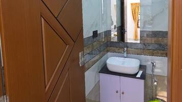 Un baño con piso de mármol, puerta de madera, lavamanos blanco y un cubo amarillo.