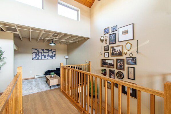 A spacious interior with a wooden staircase, a ceiling with exposed beams, and a wall adorned with framed pictures and a mirror.