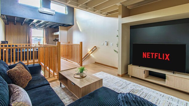 A modern living room with a large flat-screen TV displaying "NETFLIX," a wooden staircase, and a cozy seating area with a sofa and coffee table.