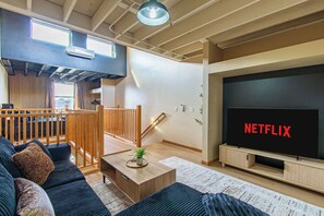 A modern living room with a large flat-screen TV displaying "NETFLIX," a wooden staircase, and a cozy seating area with a sofa and coffee table.