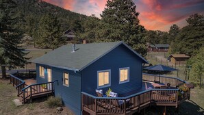 A blue house with a deck, surrounded by trees and a sunset sky. - Whispering Pines Getaway with hot tub, large deck, game room and wildlife views (Estes Park)