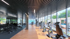 Academia com equipamentos de ginástica, amplas janelas e piso de madeira.
