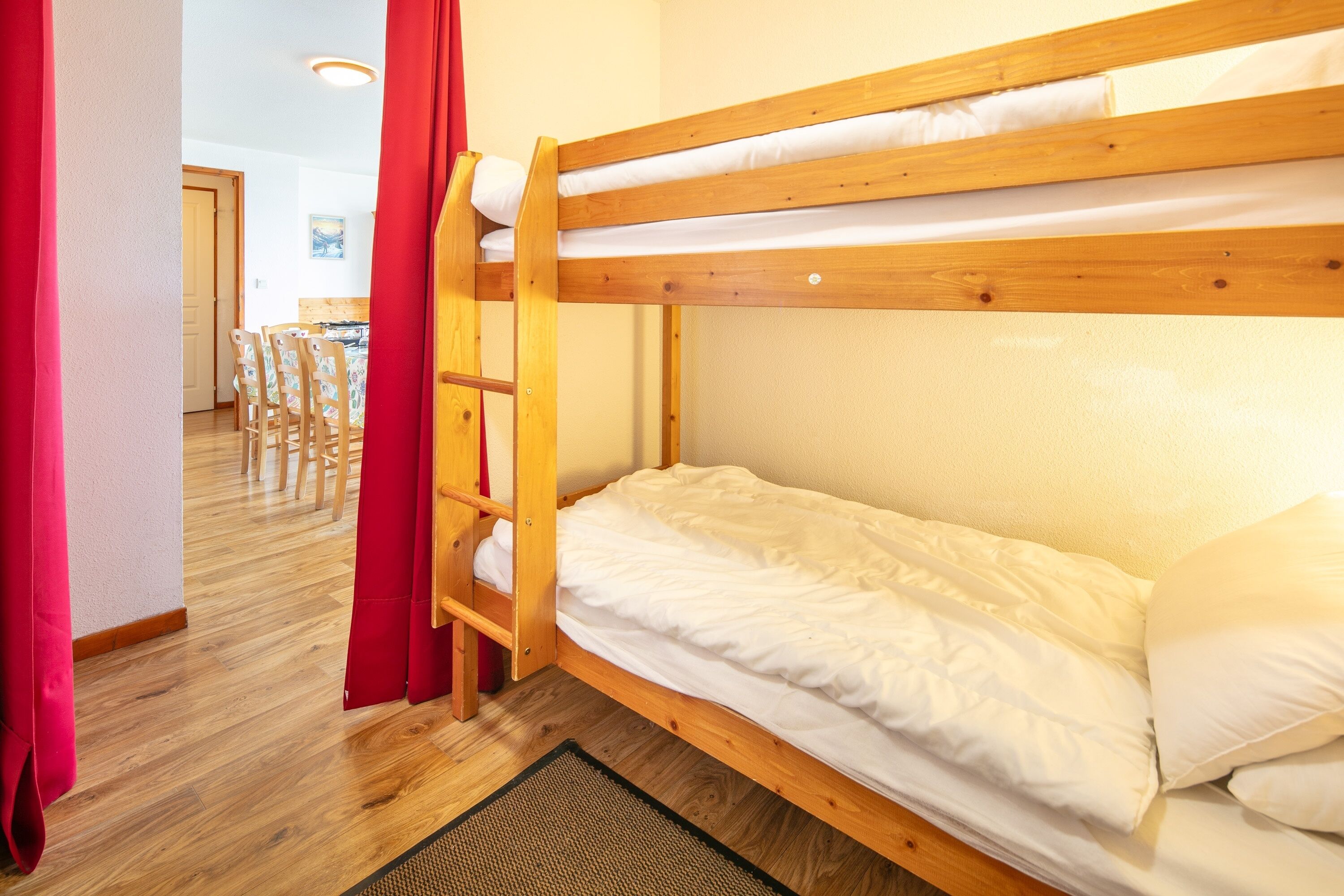 A bunk bed with a wooden frame and white bedding, a red curtain, and a dining area with chairs and a table in the background.