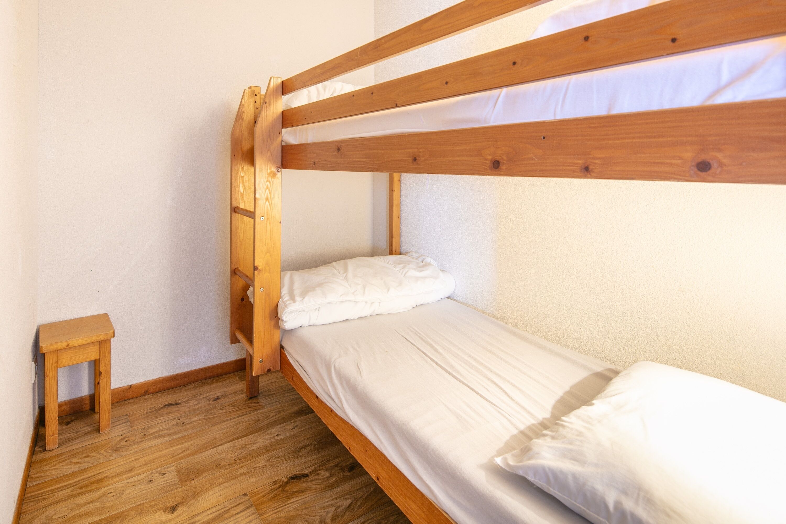 Uma cama beliche com estrutura de madeira e roupa de cama branca num quarto com soalho de madeira e um pequeno banquinho de madeira.