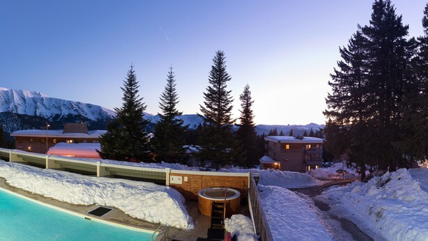 Zona de piscina exterior cubierta de nieve con jacuzzi, rodeada de edificios y montañas nevadas al fondo.