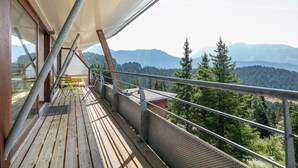 Terraza de madera con barandilla de metal que da a una cadena montañosa boscosa.
