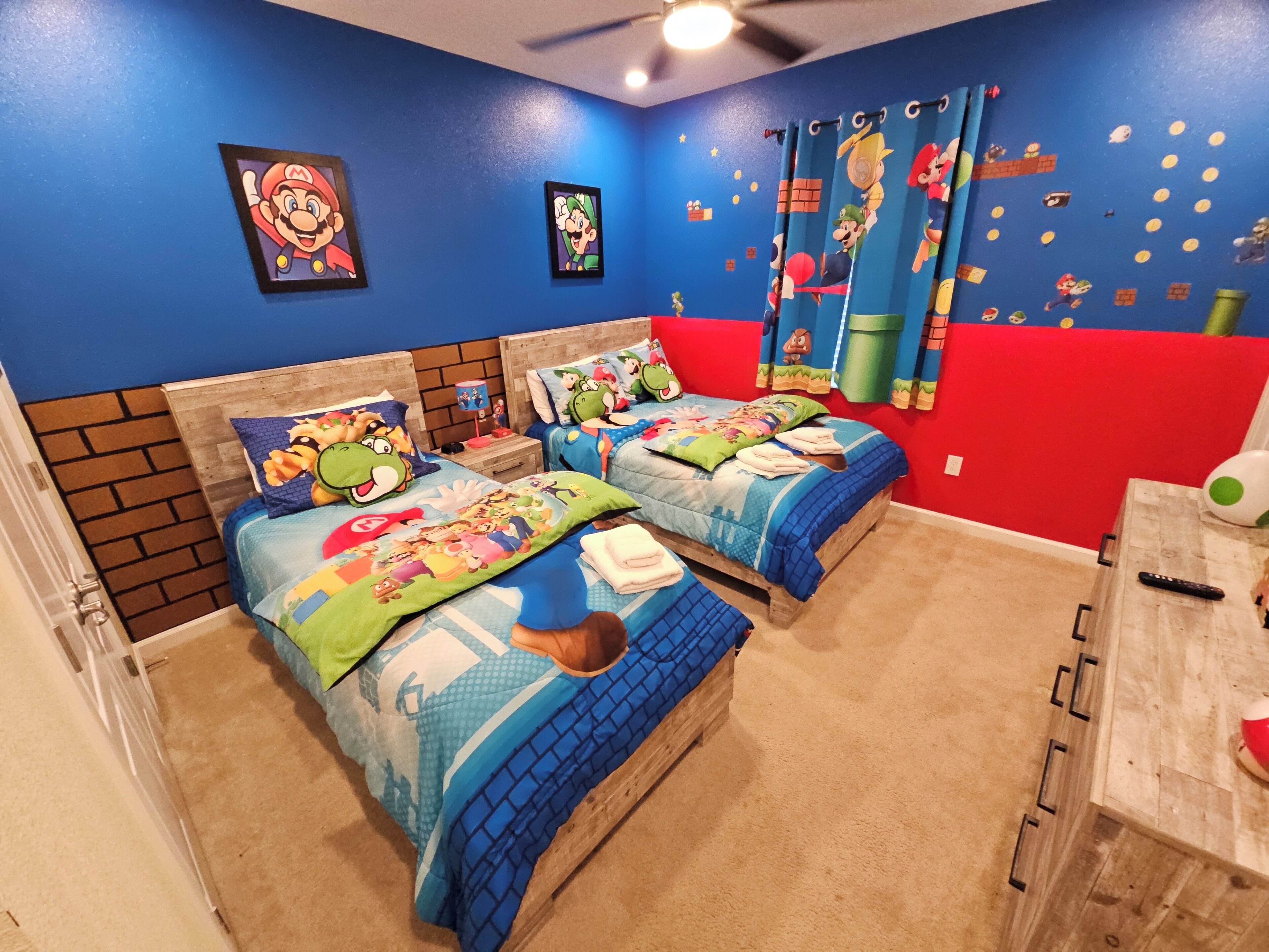 Une chambre avec deux lits, chacun agrémenté de draps et d’oreillers aux couleurs de Mario. Les murs de la chambre sont bleus, et un mur est peint en rouge pour créer un effet de contraste.