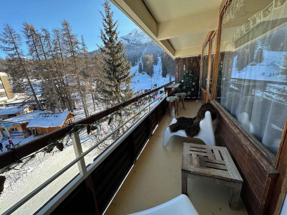 En balkon med udsigt til et snækkedækket bjerglandskab, en træbænk og en stol.