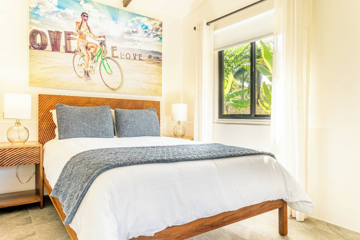 Un dormitorio con cama de madera, dos almohadas, una mesita de noche con lámpara, una ventana con cortinas y un cartel en la pared.