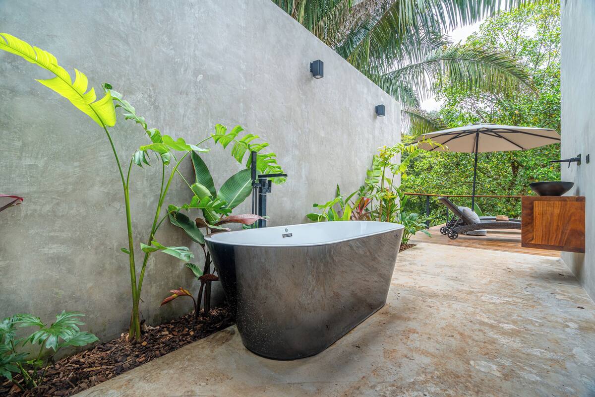 Un baño exterior moderno con una bañera grande, rodeado de abundante vegetación y un muro de hormigón.