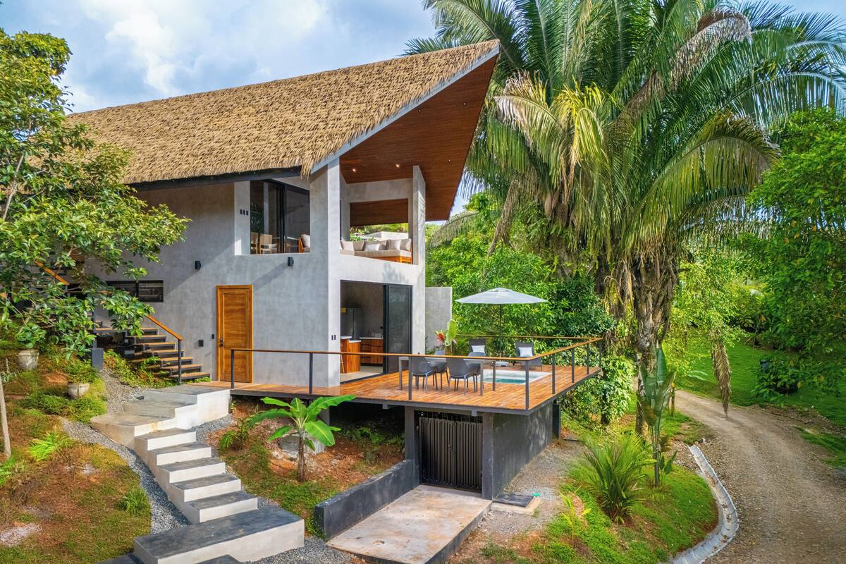 Una casa moderna con terraza de madera, una escalera que conduce a un segundo piso y un jardín exuberante con palmeras.