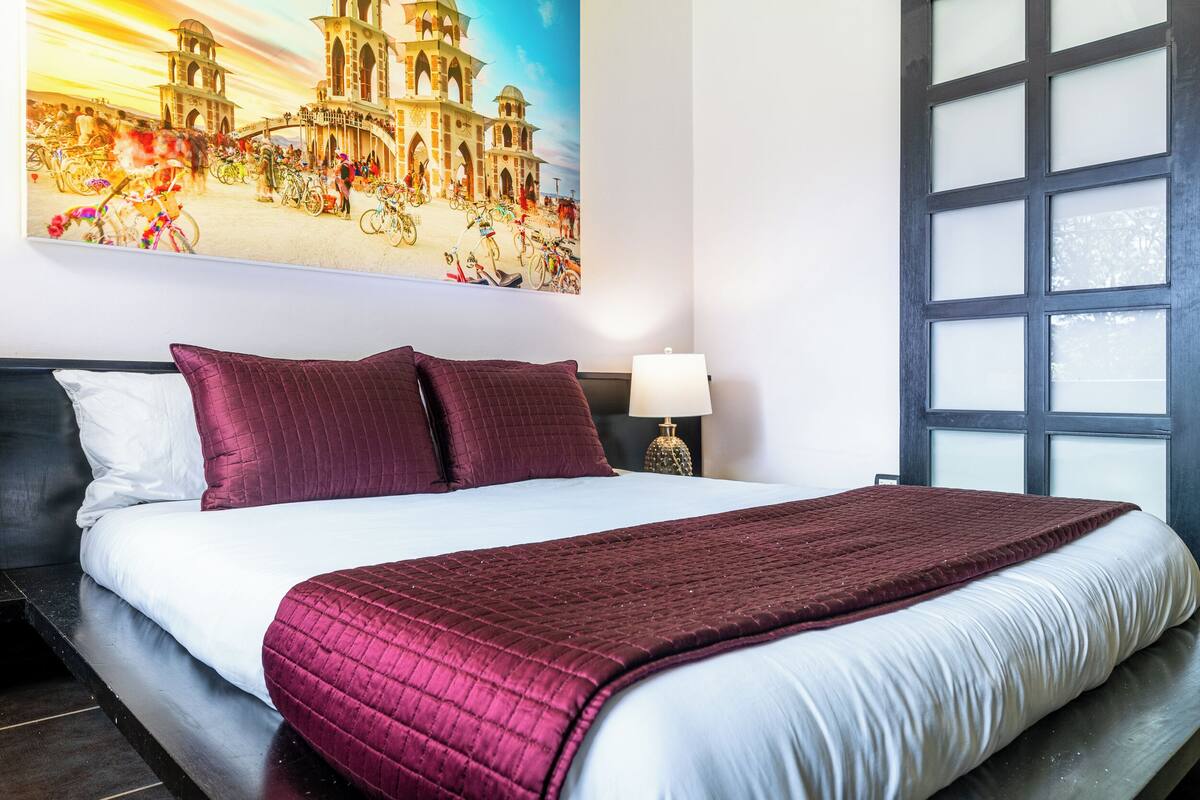 Habitación de hotel con una cama grande, una mesita de noche con lámpara, una ventana con vidrio esmerilado y un mural de un edificio histórico en la pared.