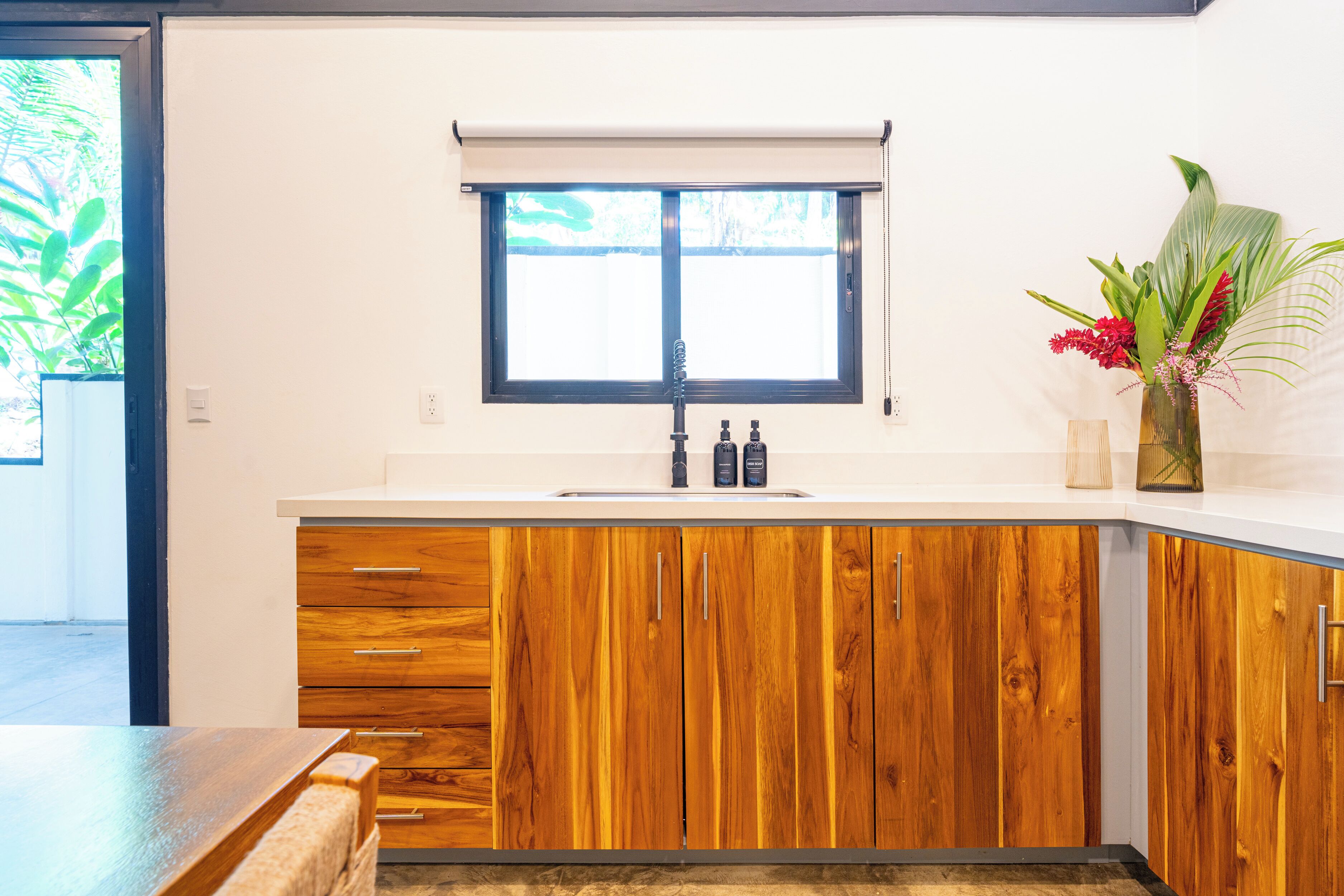 Une cuisine avec des meubles de rangement en bois, un plan de travail blanc, une fenêtre avec un store enrouleur et un vase rempli de fleurs posé sur le plan de travail.