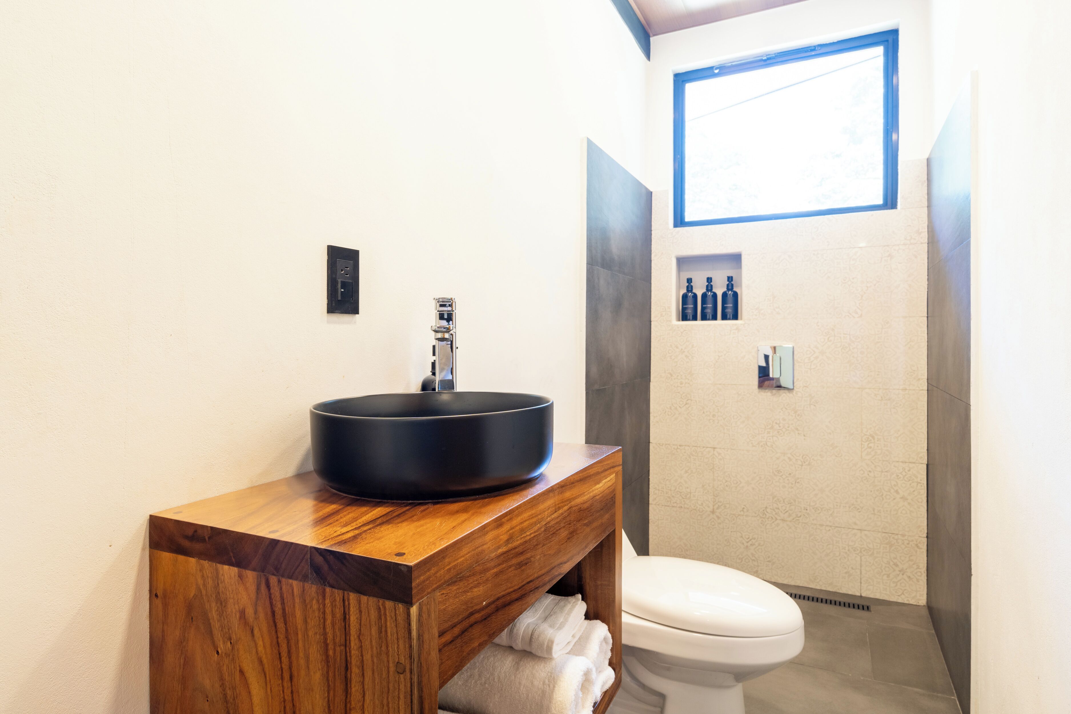 A bathroom with a wooden vanity, a black sink, a toilet, and a shower area.