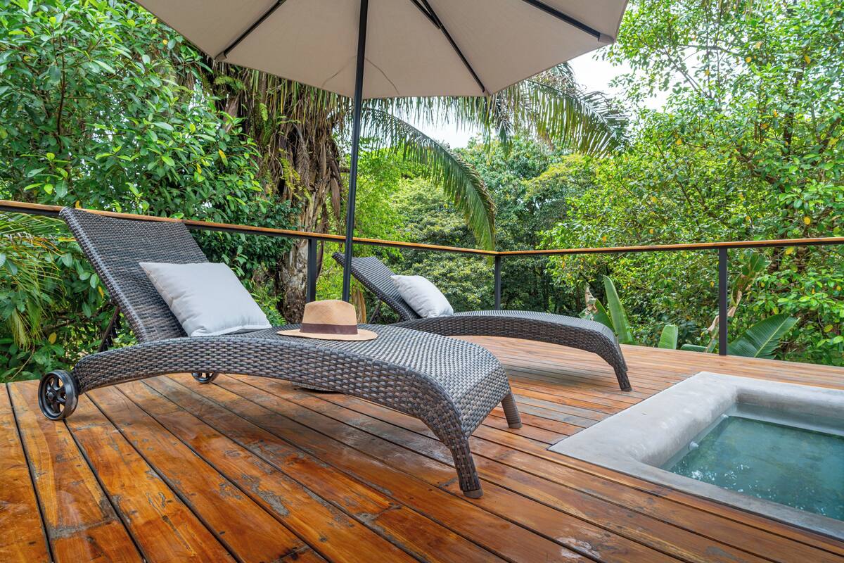 Una terraza de madera con dos sillones de mimbre, una piscina pequeña y exuberante vegetación al fondo.
