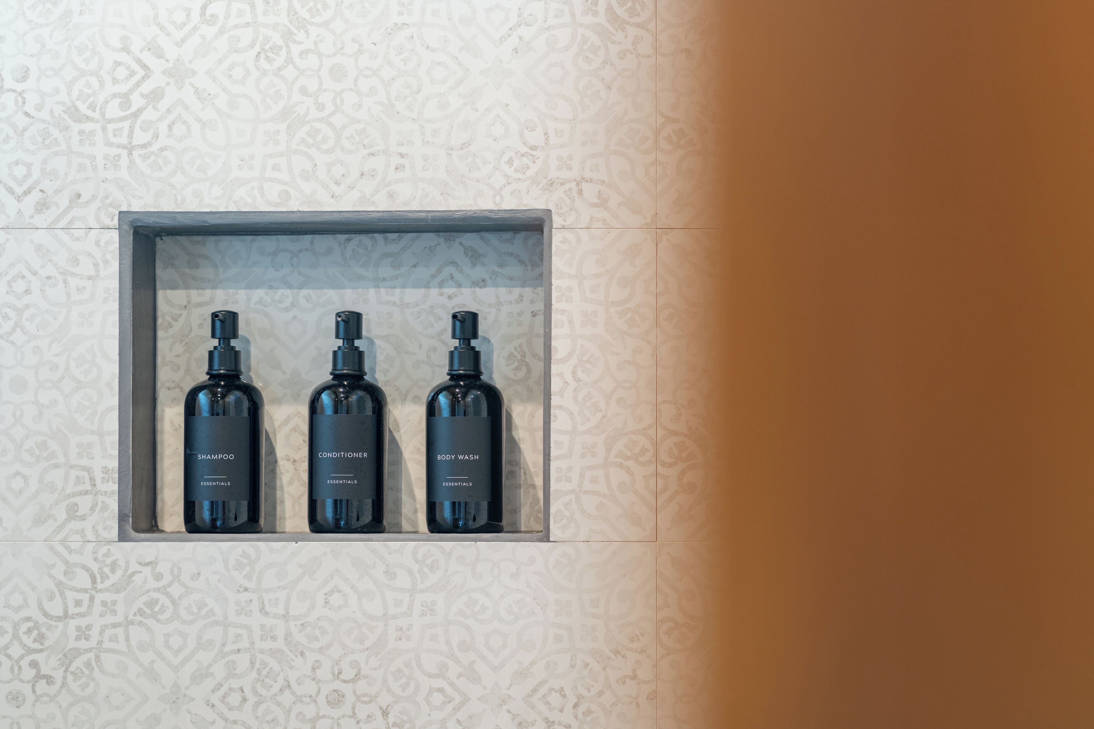 A bathroom with a patterned tile wall and a shelf containing three bottles of shampoo, conditioner, and body wash.