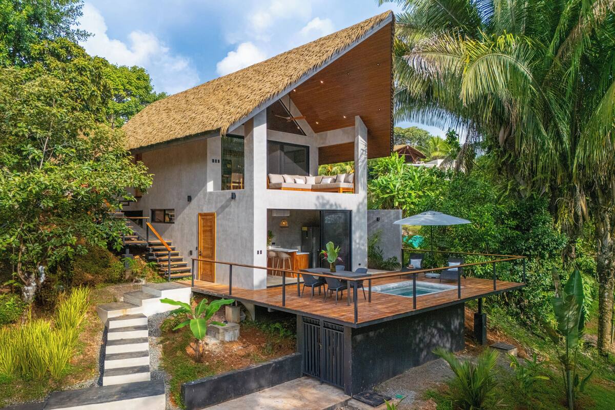 Una casa moderna de dos pisos con techo de paja, terraza de madera y piscina.