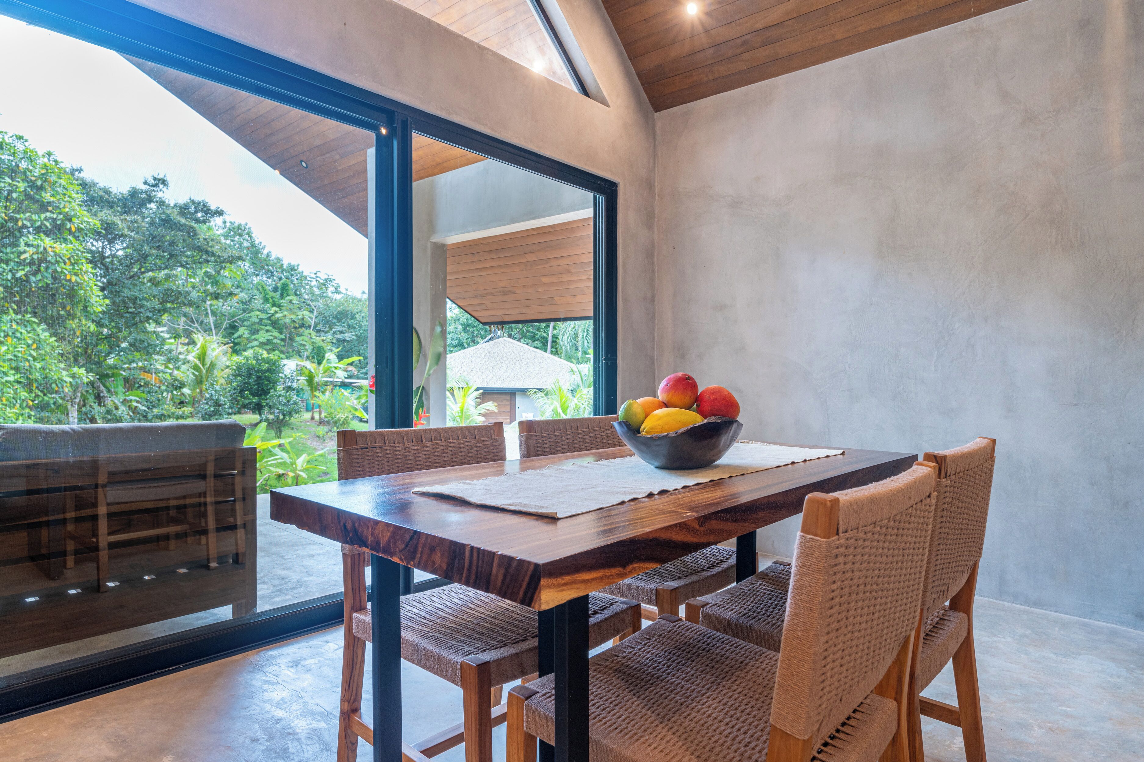 A dining area with a wooden table, wicker chairs, and a bowl of fruit.