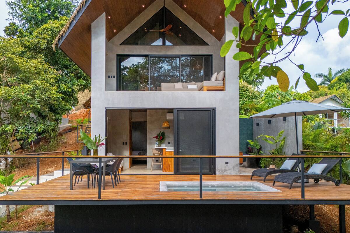 Una casa moderna de dos pisos con terraza de madera, mobiliario exterior y jacuzzi.