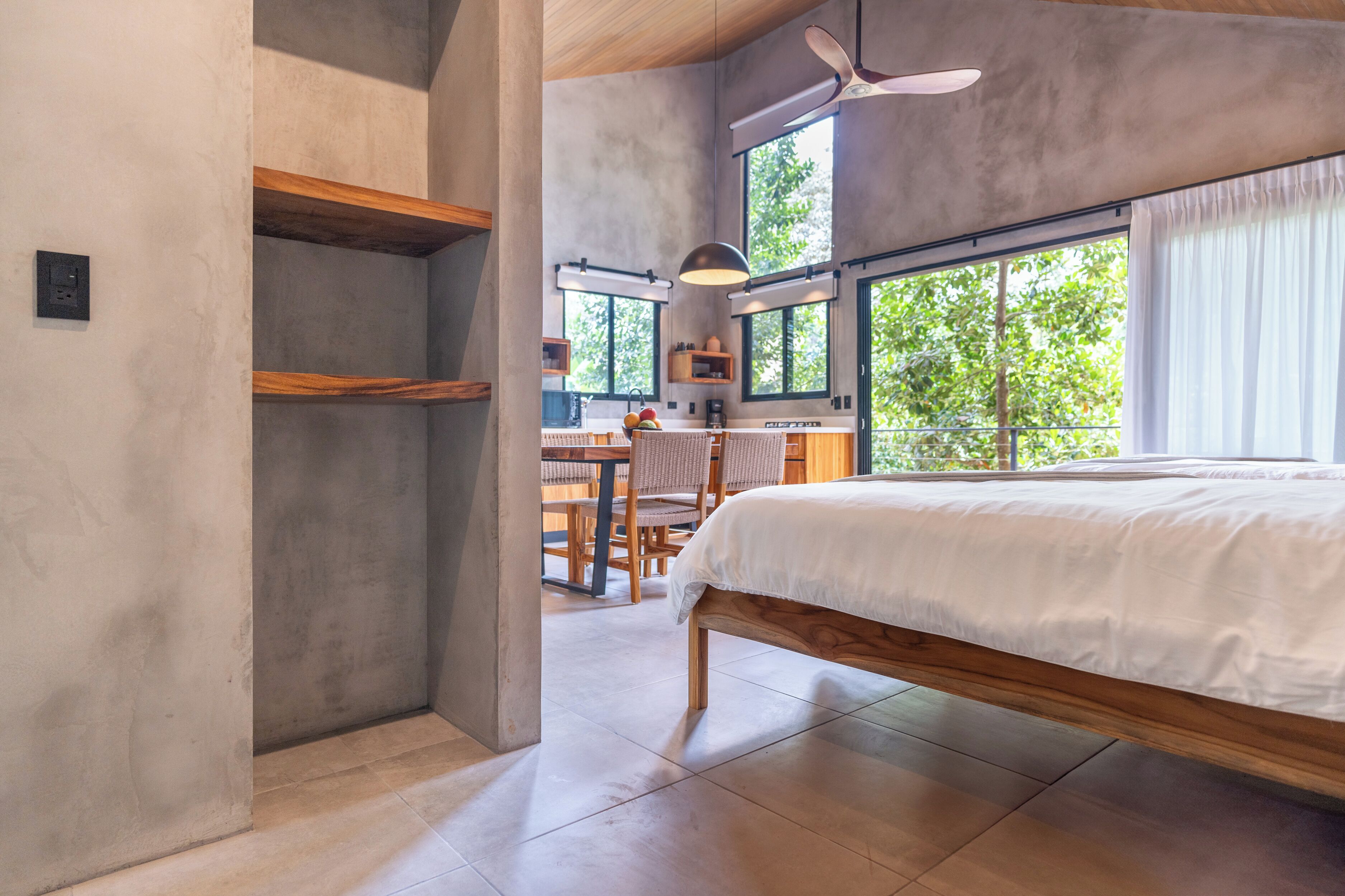 A bedroom with a large bed, wooden beams, a dining area, and a view of greenery outside.