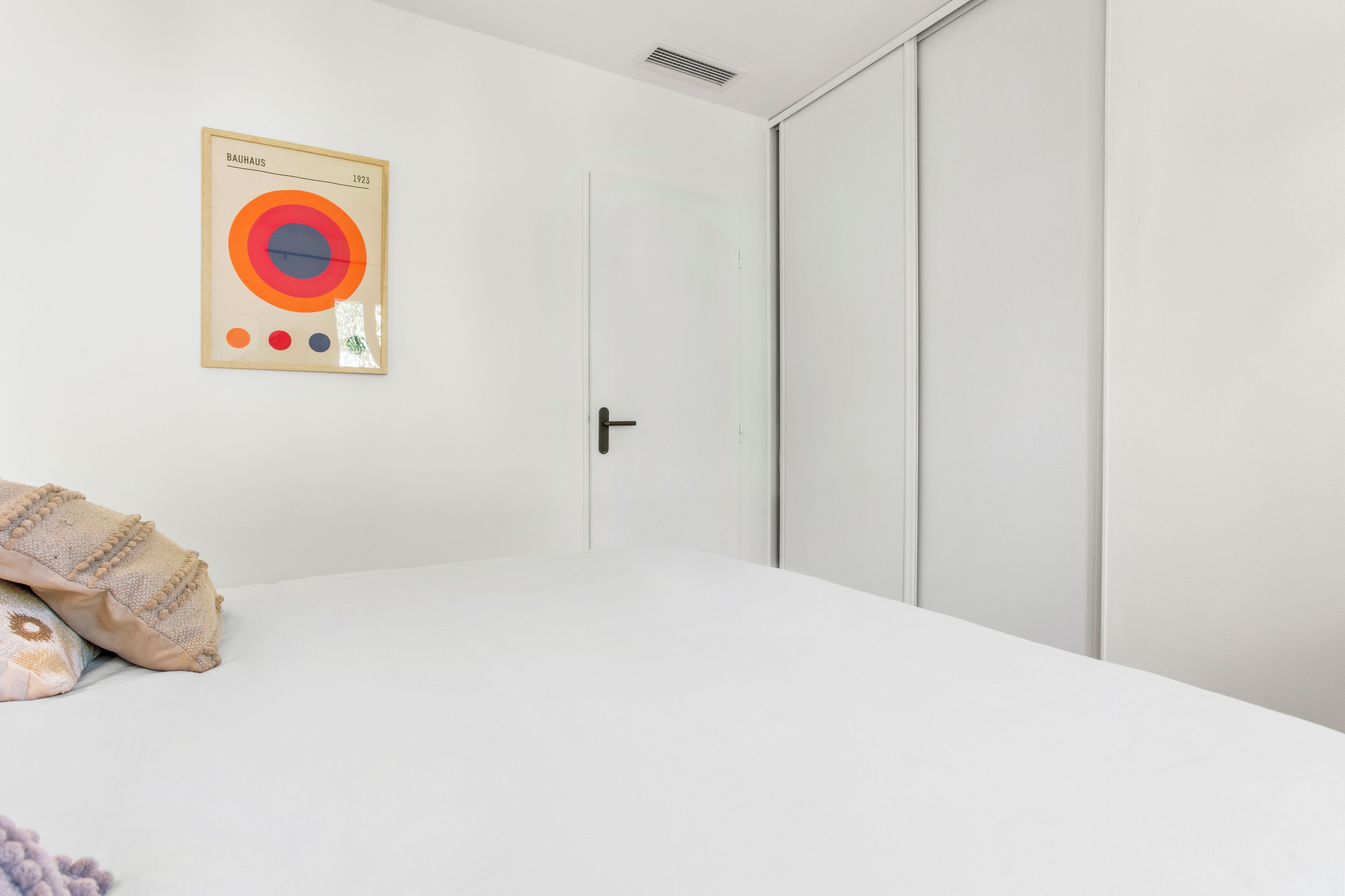 Ett minimalistiskt sovrum med en säng, en dörr och ett konstverk i Bauhaus-stil på väggen.