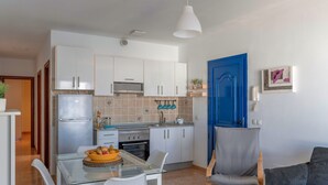 Una cocina compacta con armarios blancos, una puerta azul y una zona de comedor con mesa y sillas de vidrio.