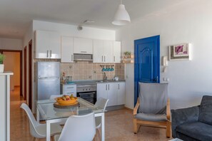 Une cuisine compacte avec des meubles blancs, une porte bleue et un coin repas comprenant une table et des chaises en verre.