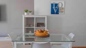 Un comedor moderno con una mesa de vidrio, sillas blancas, un cuenco de fruta, una pequeña planta en maceta y un cartel que dice 'Home' en un mueble blanco.