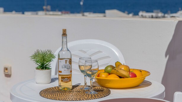 Una mesa con una botella, copas, un cuenco de fruta y una planta en maceta en un balcón con vistas al mar.