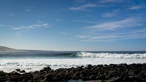 Una costa rocosa con olas, cielo despejado y colinas al fondo.