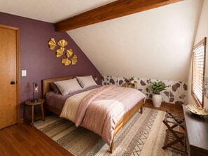 A bedroom with a sloped ceiling, a bed with a pink bedspread, a round nightstand with a lamp, a potted plant, and a decorative wall piece.