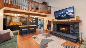A spacious living area with a fireplace, a television mounted above it, a kitchen in the background, and a balcony with wooden railings.
