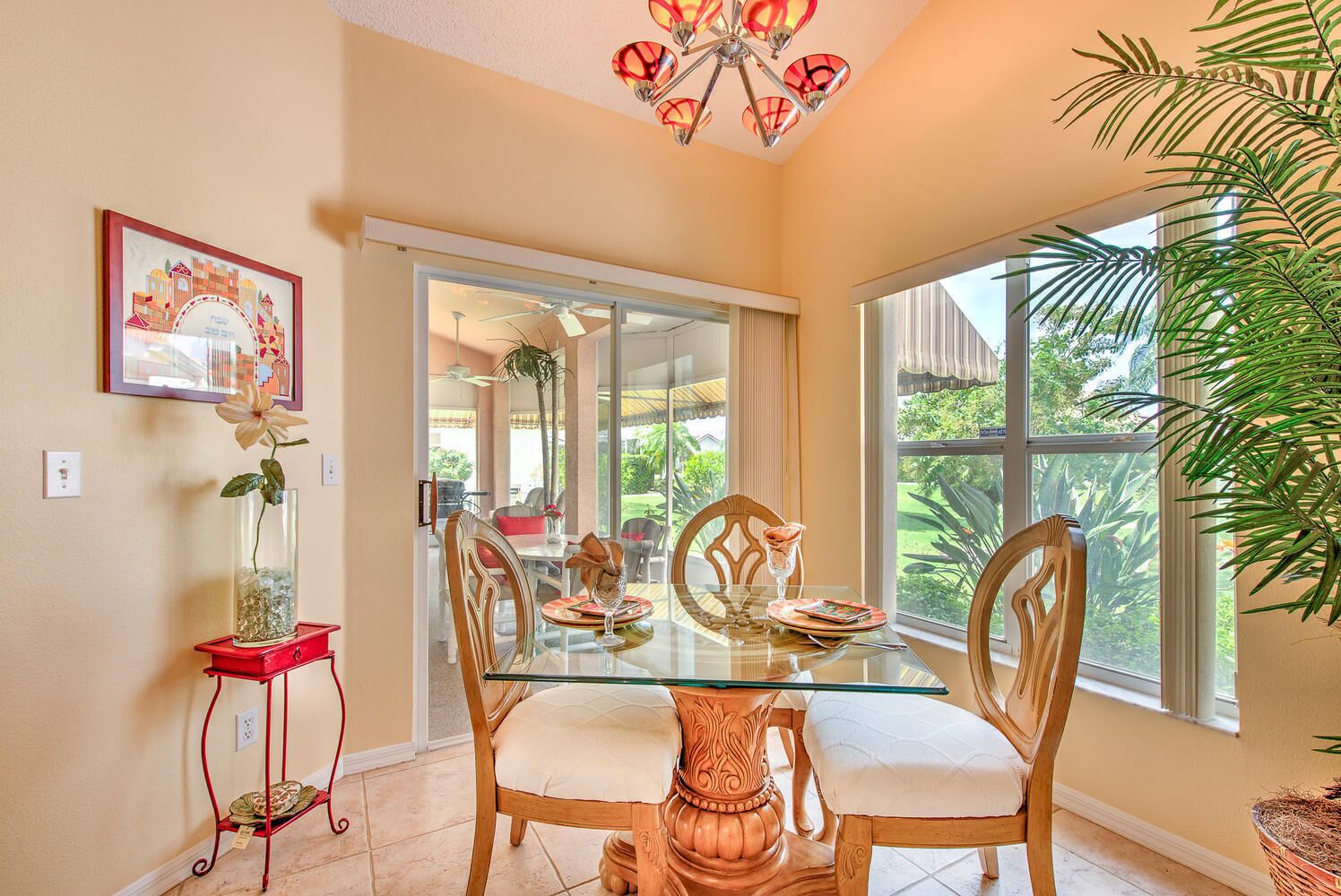 A dining area with a glass table, wooden chairs, a red side table, and a large window offering a view of greenery.