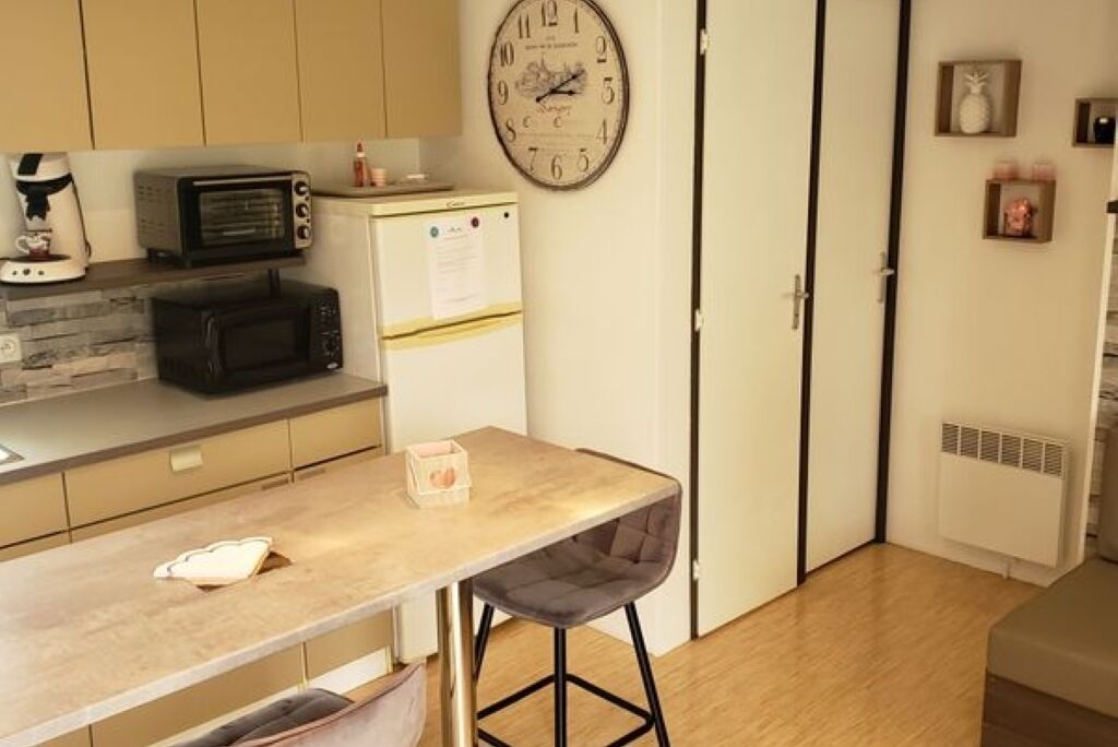 A compact kitchen with a microwave, toaster, and refrigerator. A clock is on the wall, and a small table with a chair is in the center.