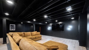 A modern living room with a sectional sofa, a flat-screen TV mounted on the wall, and recessed lighting. - Modern 4BR Home on Golf Course + Sauna Close to Campus, Dining & Entertainment (Greencastle)