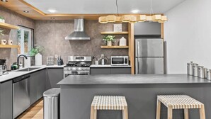 Une cuisine moderne dotée d’un îlot central, d’appareils électroménagers en acier inoxydable, d’étagères en bois et d’un luminaire suspendu.