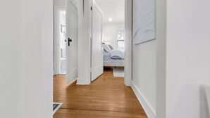 A hallway with wooden flooring, white walls, and a bed in the adjacent room.
