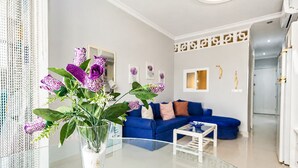 A living room with a blue sofa, a glass coffee table, and a vase with purple flowers on a glass table.