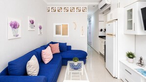 A modern living room with a blue sofa, white cabinets, and a kitchen area with white appliances.